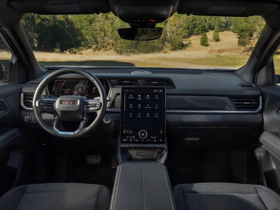 Interior dashboard view of a GMC SUV with a portrait touchscreen display and wood-trimmed dash, looking out over a gravel road and green field