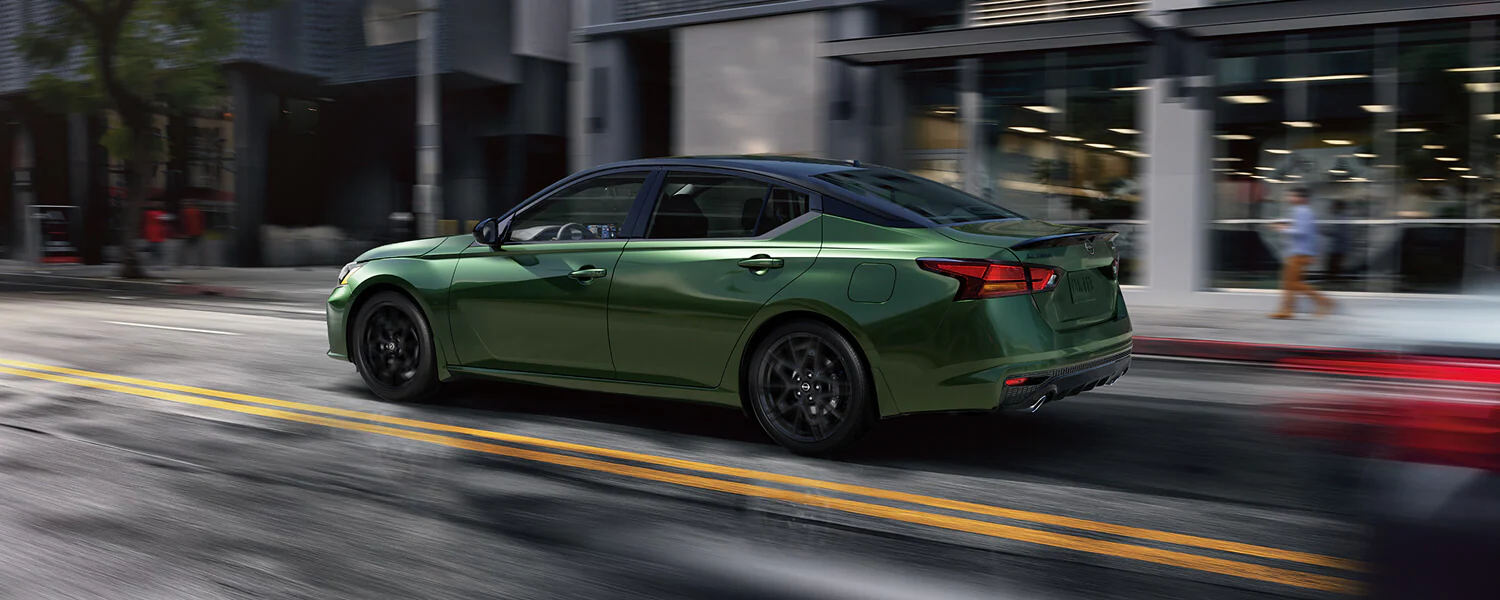 Green Nissan Altima SR sedan with a black roof and dark alloy wheels driving on a city street, shown from a rear three-quarter angle with motion-blurred surroundings.