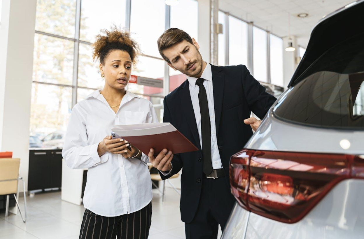 A female customer holding a folder and a male car salesman discussing a silver car in a dealership.
