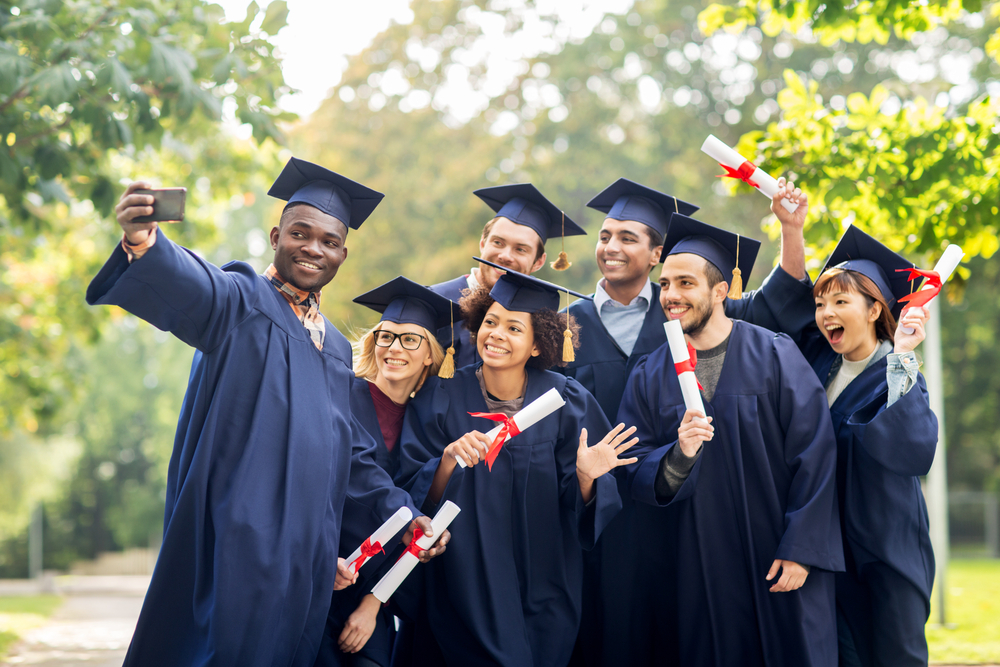 A Group of college graduates  all dressed up in their cap and gown regalia and all holding their diplomas, one person is holding their phone out for a selfie  and everyone is smiling