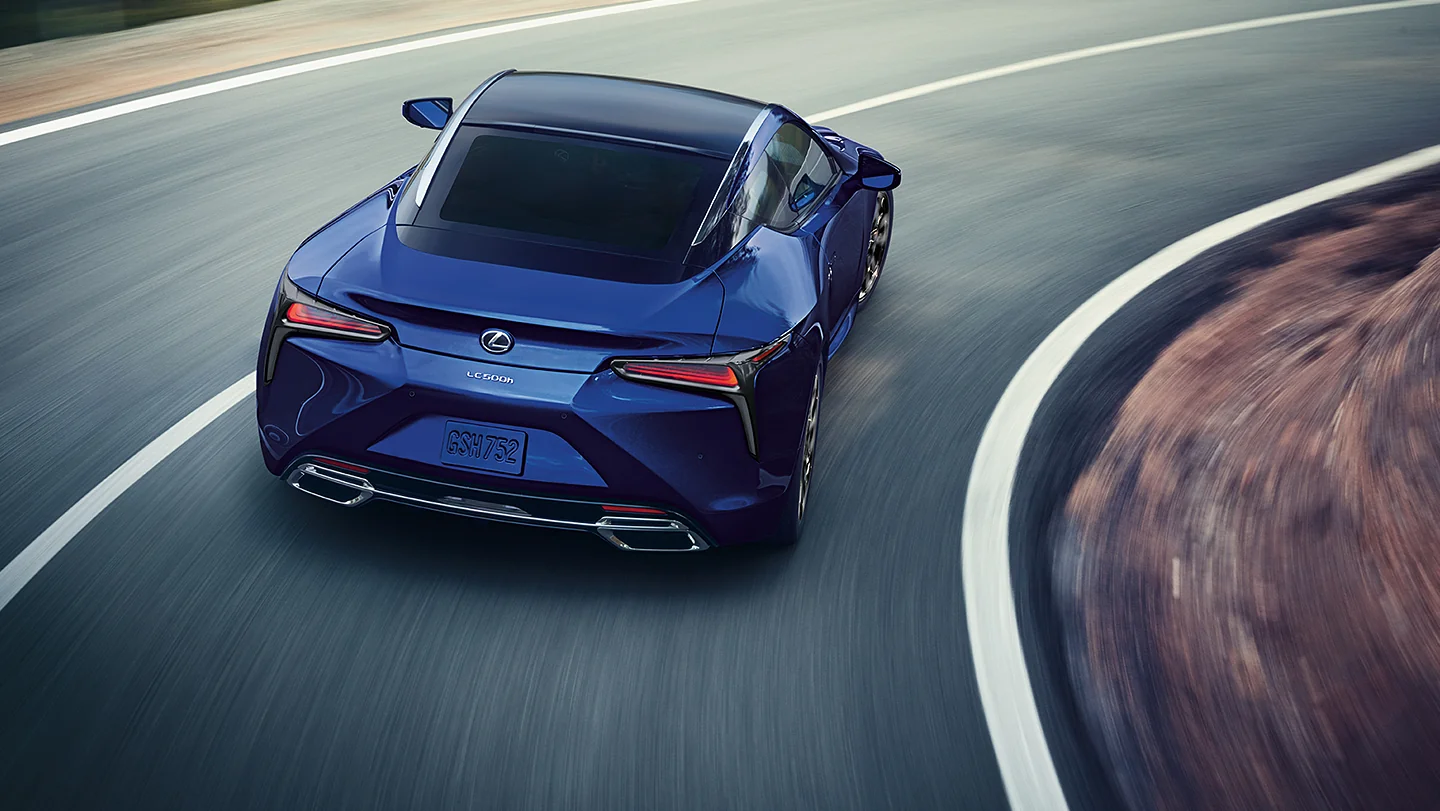 A blue Lexus LC 500h coupe driving with motion blur around a curving road, shown from an elevated rear three-quarter angle.