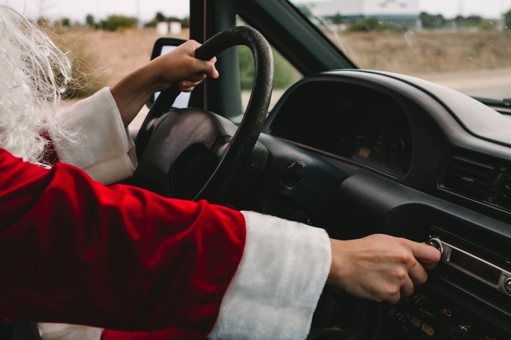 Santa Claus reaches to turn a dial on his radio inside of a car while he's driving