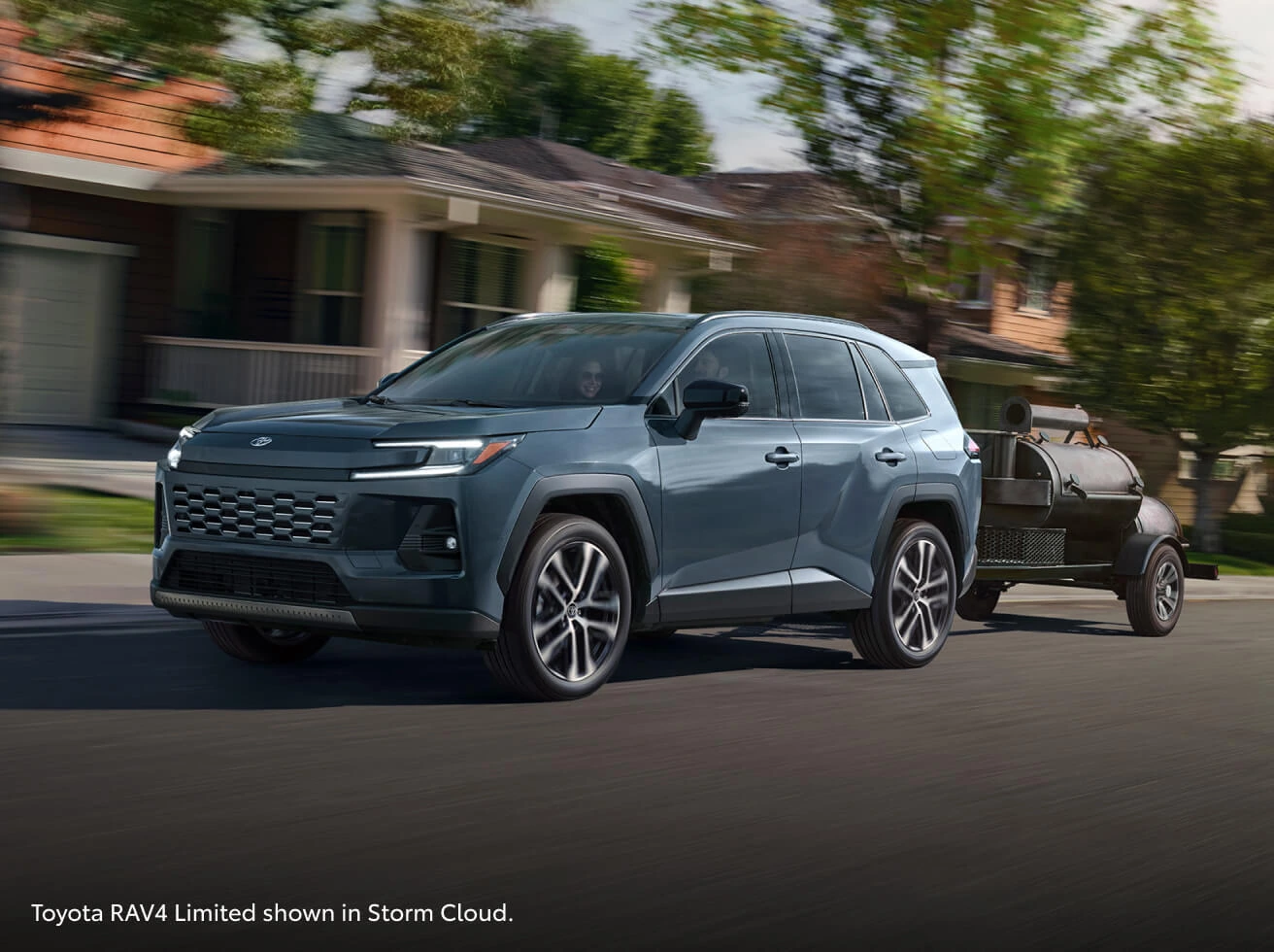 Front three-quarter view of a 2026 Toyota RAV4 Limited in Storm Cloud towing a smoker trailer through a suburban neighborhood.