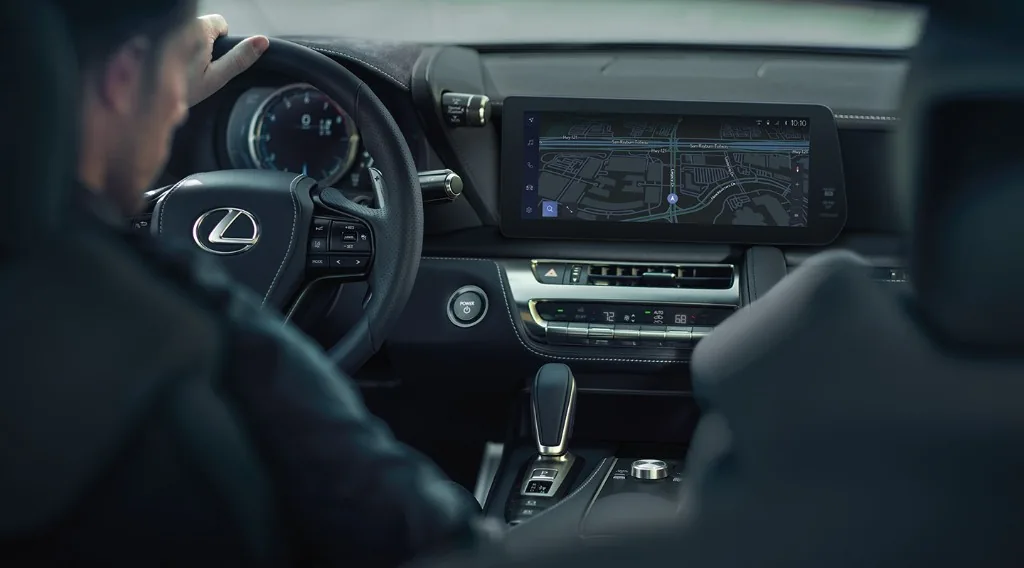 View from behind the driver showing a Lexus steering wheel and headrest with the Lexus logo, an infotainment screen displaying a navigation map, and the center console with climate controls and gear shifter.