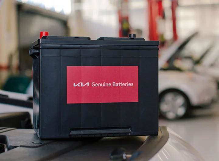 A Kia Genuine Batteries battery sitting on a vehicle's roof in a service bay, with another car's open hood visible blurred in the background.
