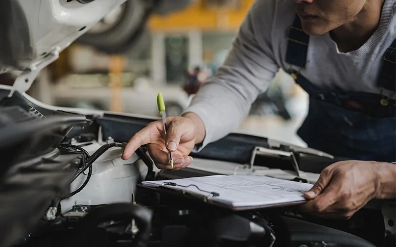 Mechanic in overalls inspecting a car engine while holding a clipboard.