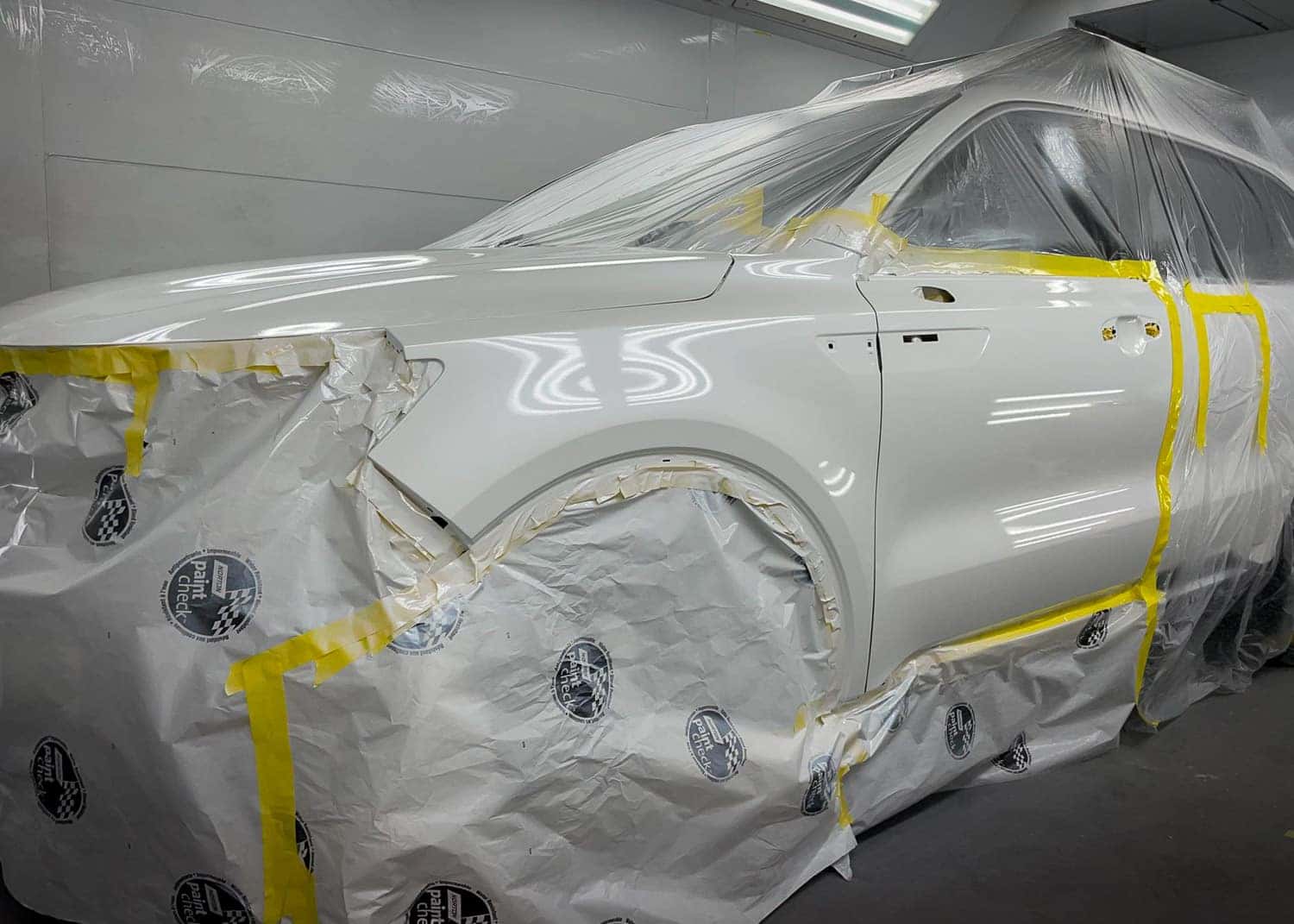 White vehicle partially covered in protective plastic and tape, undergoing collision repair in a pro…