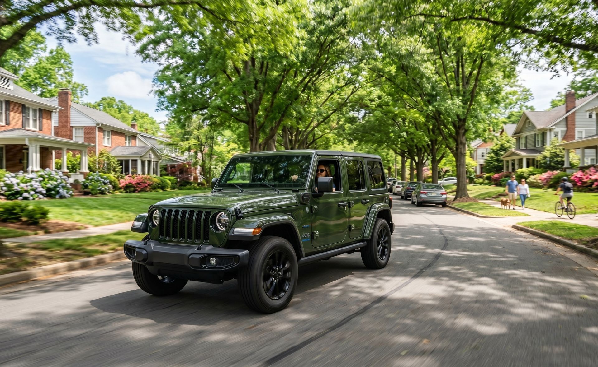 A Sarge Green Jeep Wrangler 4xe plug-in hybrid SUV driving down a beautiful, tree-lined residential neighborhood street past classic brick houses and manicured lawns.