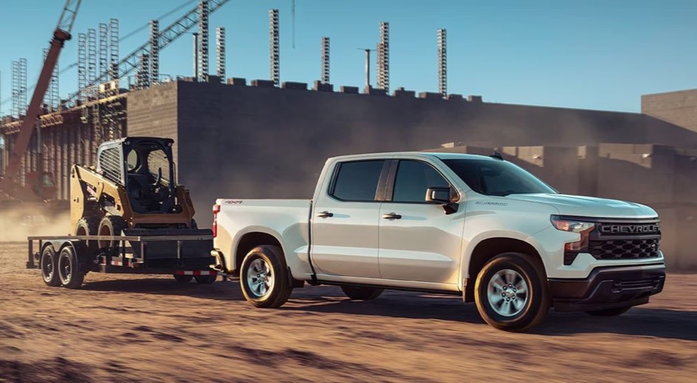 A white 2023 Chevy Silverado 1500 WT is shown from the side while towing a skid-steer.