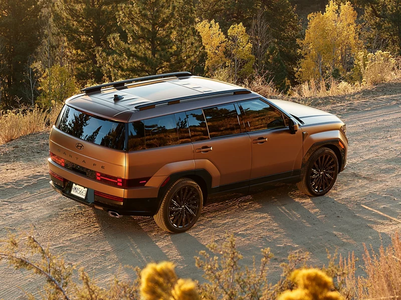 Copper 2027 Hyundai Santa Fe SUV with black wheels and roof rails, seen from above at the rear three-quarter on a sandy dirt road lined with golden autumn trees and dry shrubs