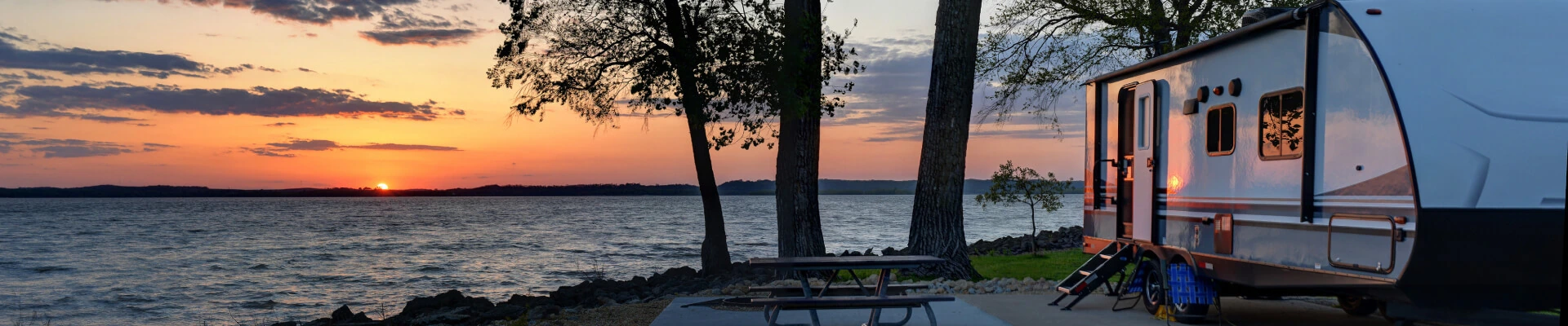 A fifth wheel camper trailer parked at a lakeside campsite at sunset.