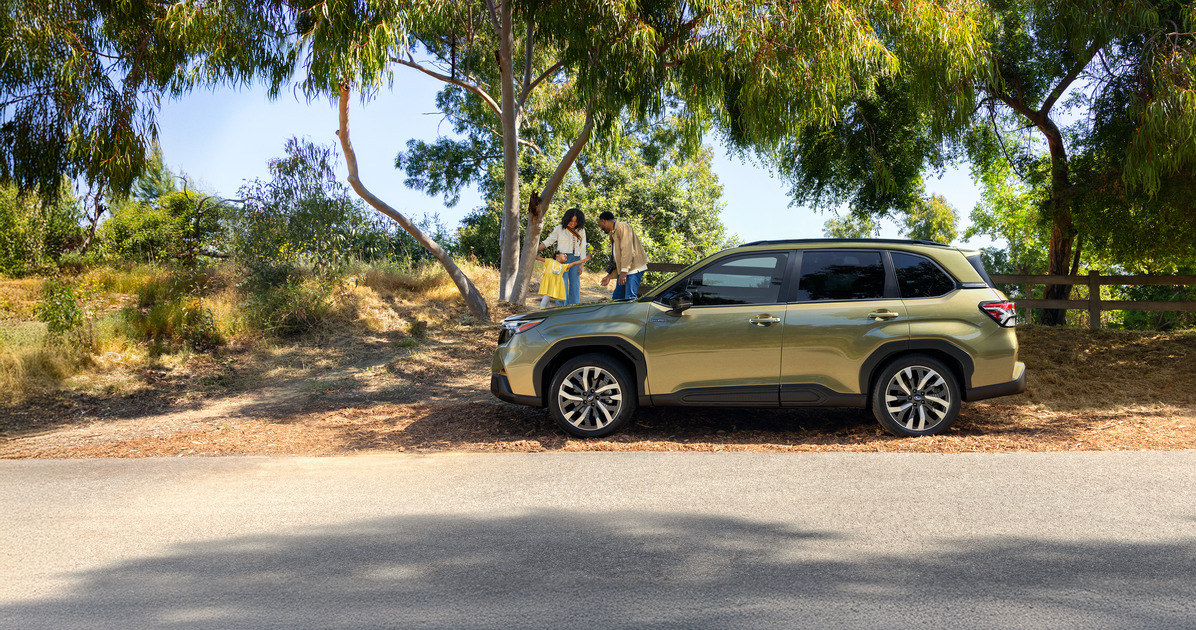 A 2026 Forester Hybrid in Autumn Green Metallic parked on the road as a family plays with their daughter in the background.