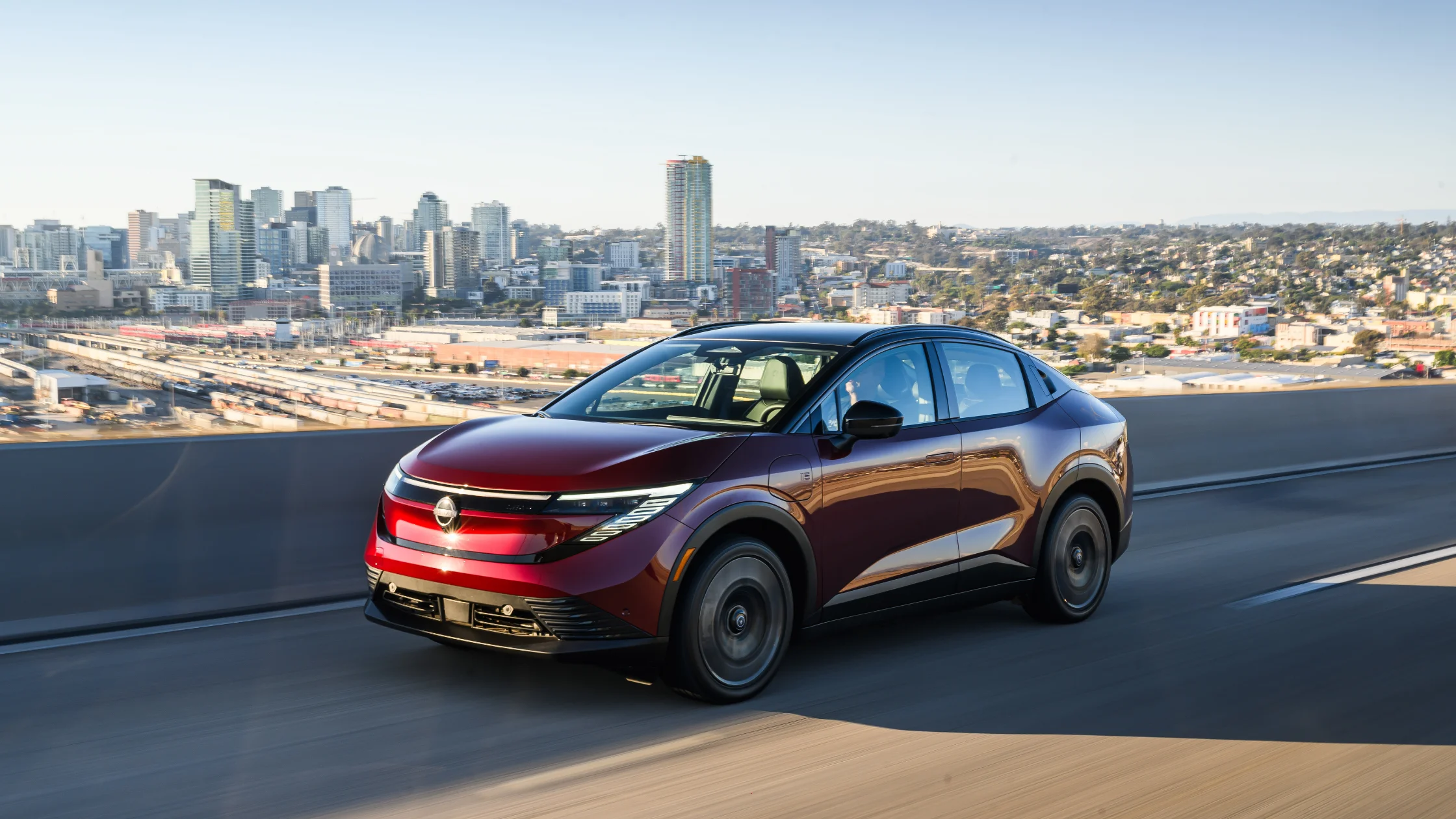 2026 Nissan LEAF® in deep red driving on an elevated city highway with a downtown skyline behind it.