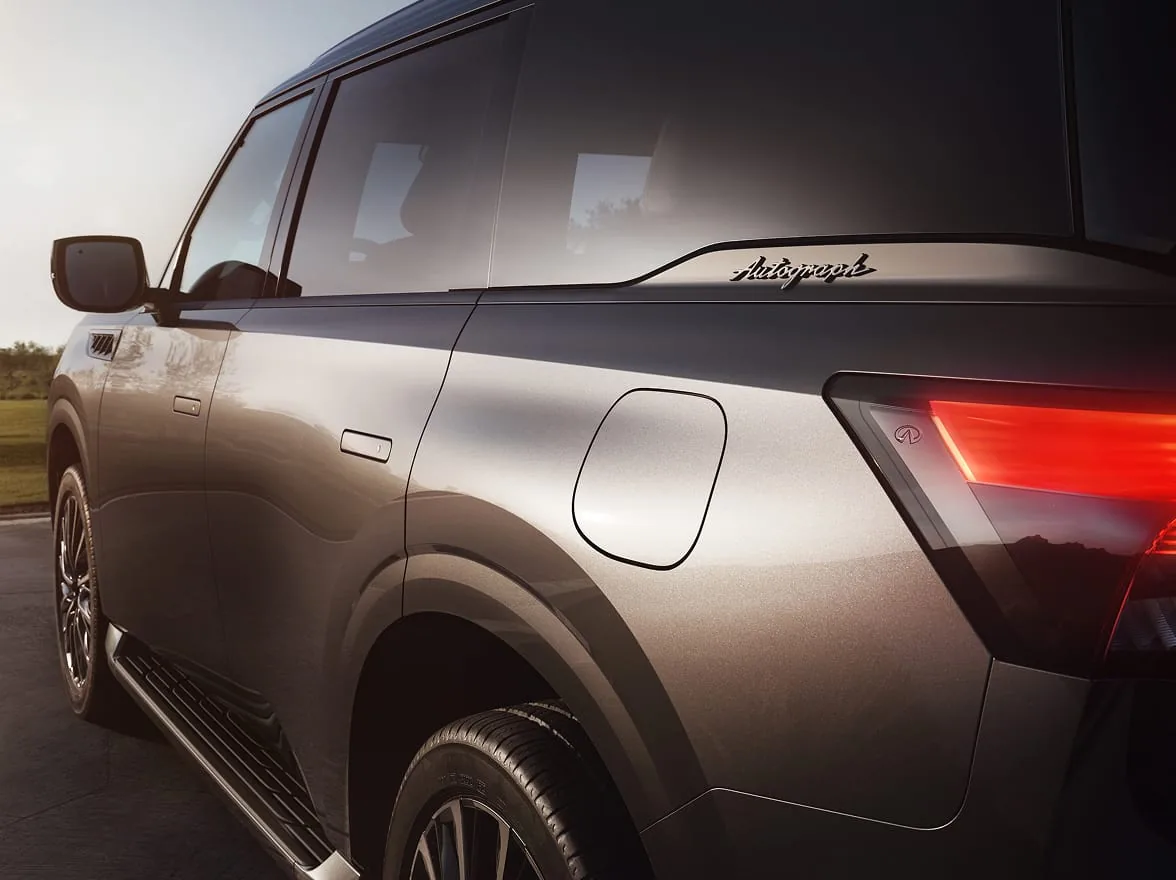 Rear three-quarter close-up of a dark metallic 2027 INFINITI QX80 AUTOGRAPH at golden hour, showing silver Autograph script on the D-pillar and a glowing segmented LED taillight.
