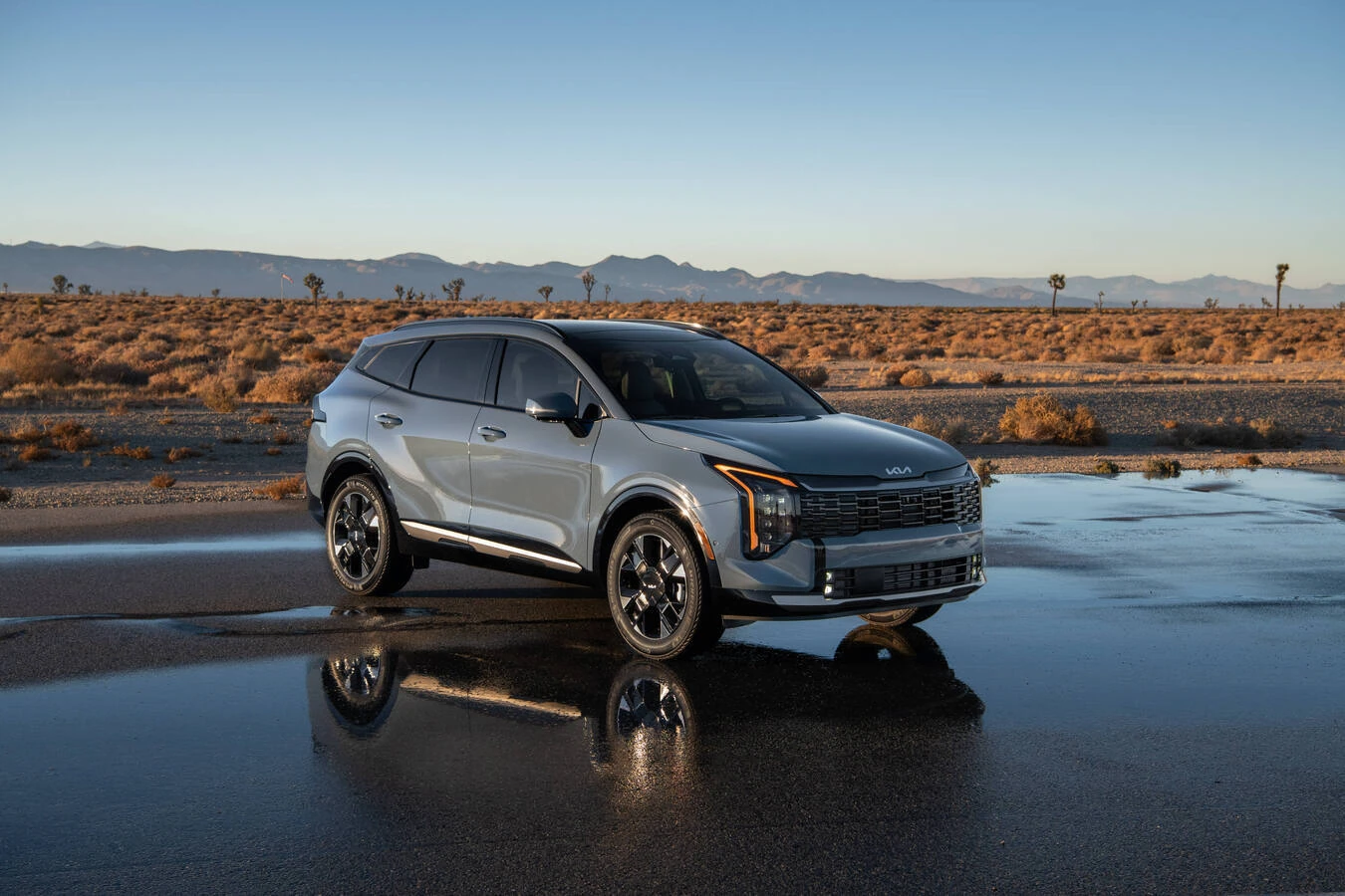 2027 Kia Sportage Hybrid parked on a wet desert road at dusk, with Joshua trees and distant mountains reflected in puddles on the pavement.