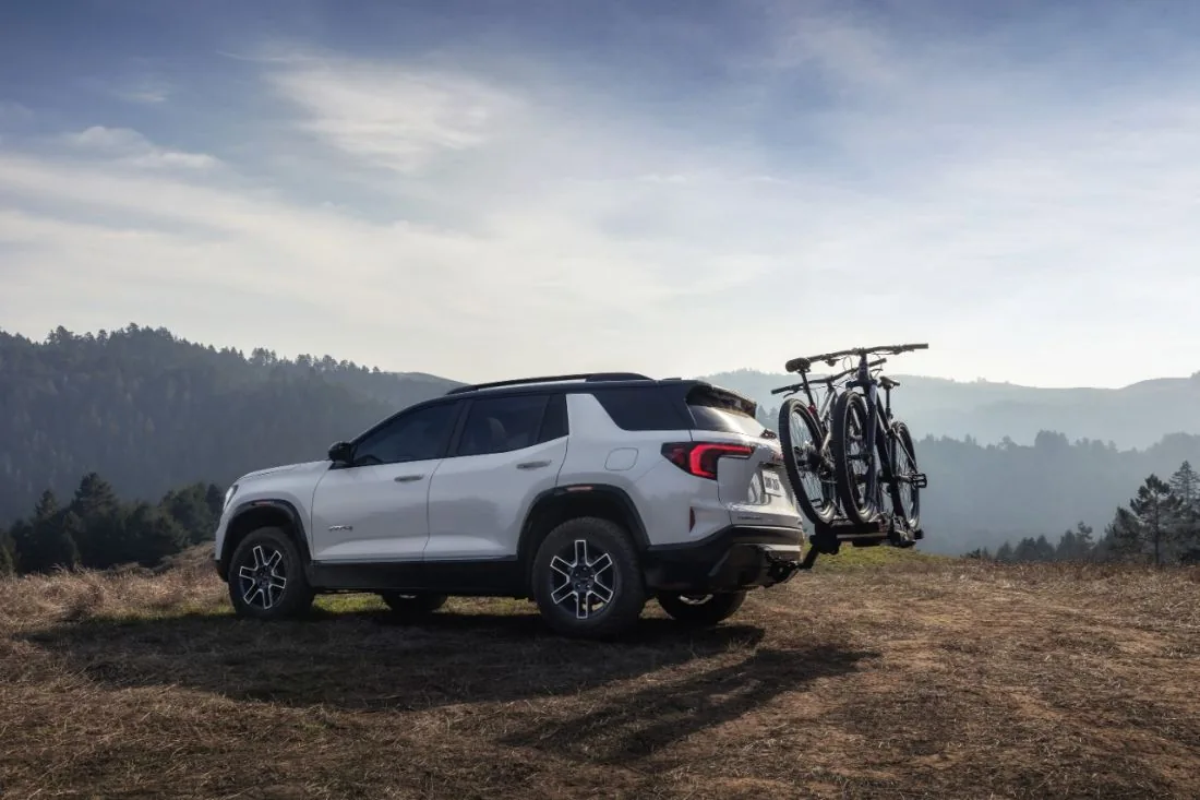 White GMC SUV with an AT4 badge and a rear-mounted bike rack carrying three bikes, parked on a hilltop with forested hills in the background