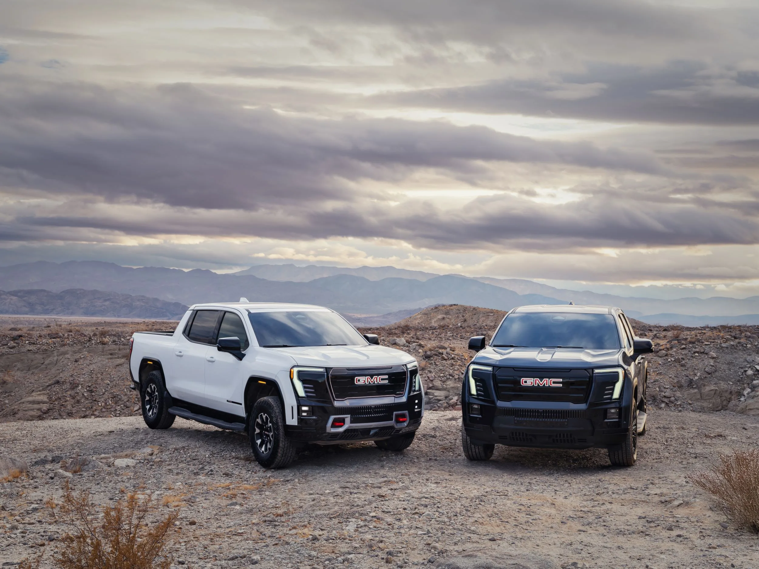 A white and a black GMC pickup truck parked side by side on a rocky desert overlook under a cloudy sky, both with a legible GMC grille badge, mountains visible in the background