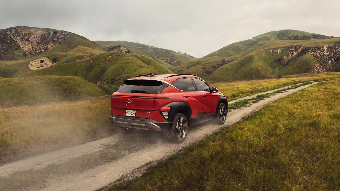 A red 2024 Kona from the rear right driving through a rolling, grassy dirt road.