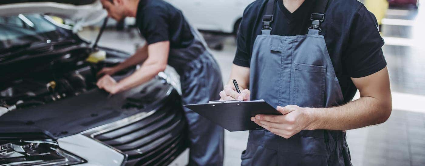 A mechanic with a clipboard is in front of another mechanic working on a vehicle.