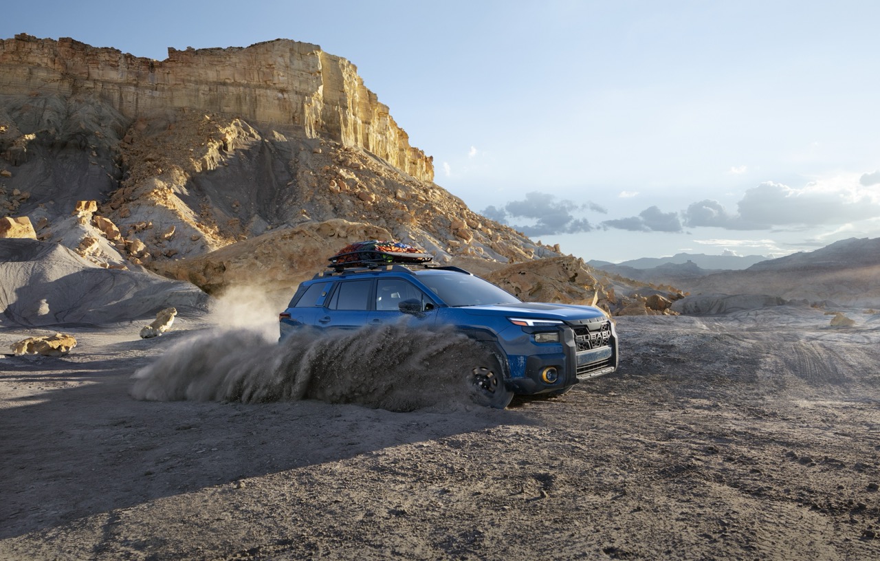 A static image of a blue 2026 Subaru Outback Wilderness driving through loose dirt in a rocky desert landscape. 
