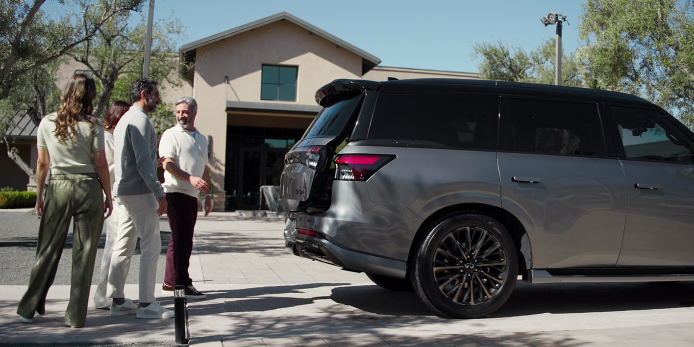 Four people stand near the open liftgate of a dark gray two-tone 2027 INFINITI QX80 parked in a sunny cobblestone driveway.