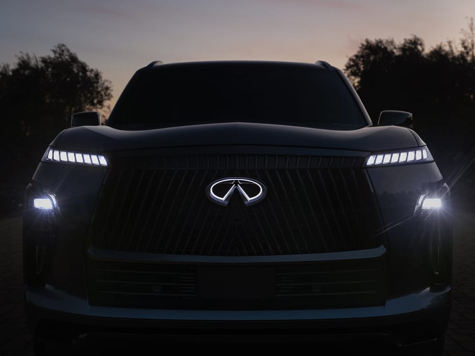 Front view of the 2027 INFINITI QX80 at dusk with an illuminated grille emblem and multi-segment LED daytime running lights glowing in low light.