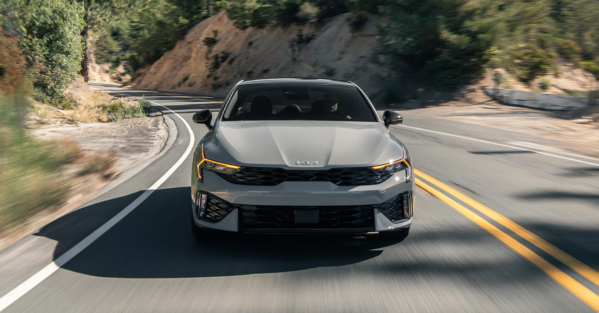 A front-facing view of a grey 2026 Kia K5 sedan driving down a winding canyon road