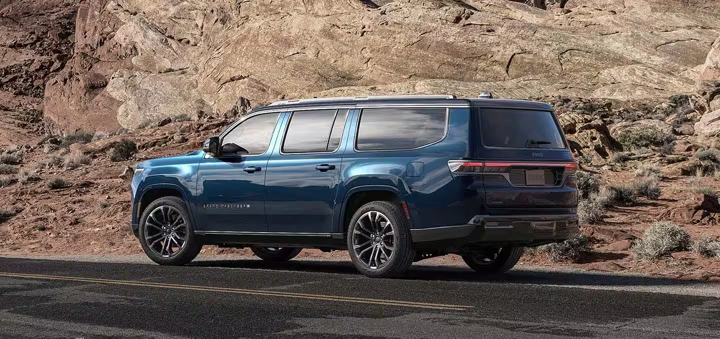 Dark blue 2027 Jeep Grand Wagoneer SUV with black multi-spoke wheels and a long body, seen from the rear three-quarter on a two-lane desert road in front of tan rock formations