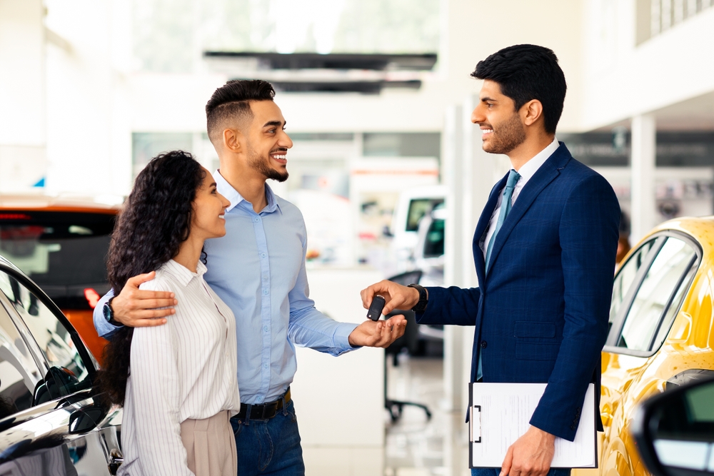 Smiling salesman in a navy blue suit hands car keys to a Hispanic couple, with the man's arm around the woman, in a car dealership showroom with vehicles in the background