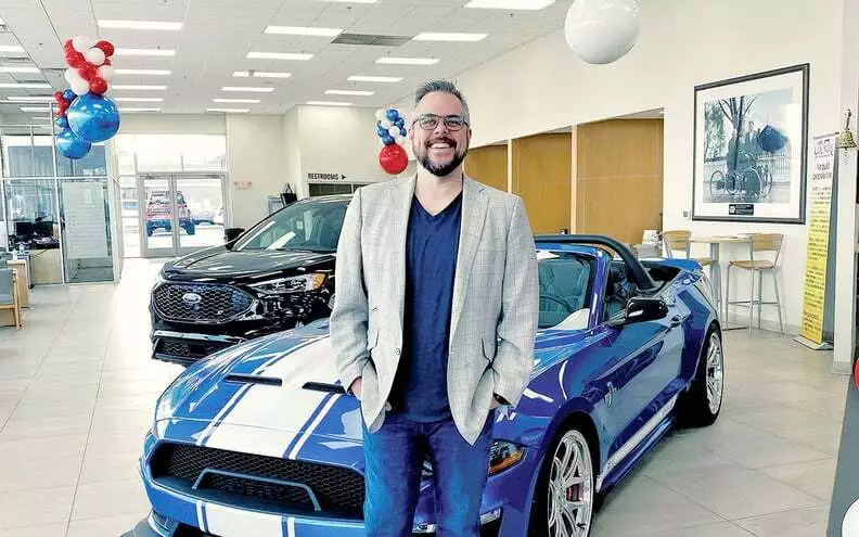 A man is standing in front of a blue car with white racing stripes and also has another black car behind the blue car, the setting is a car dealership with numerous balloons around