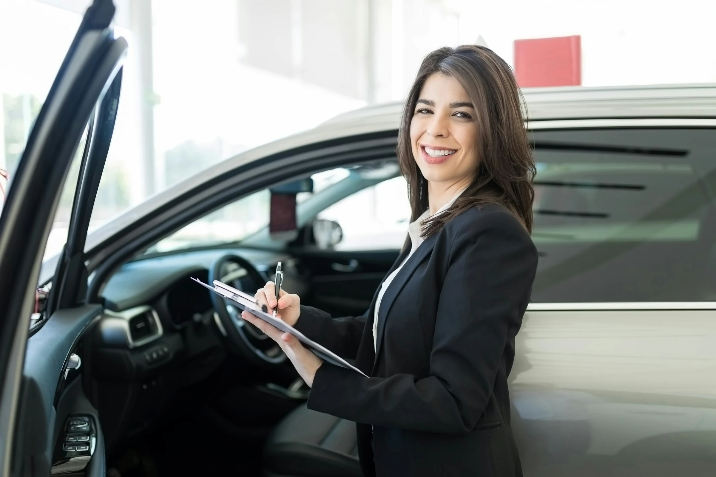 Smiling salesperson in a blazer signing paperwork on a clipboard beside an SUV with its door open in a dealership showroom