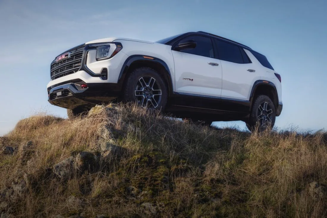 Front three-quarter view of a white GMC SUV with off-road-styled wheels and black cladding, parked atop a rocky, grassy hill under a clear sky