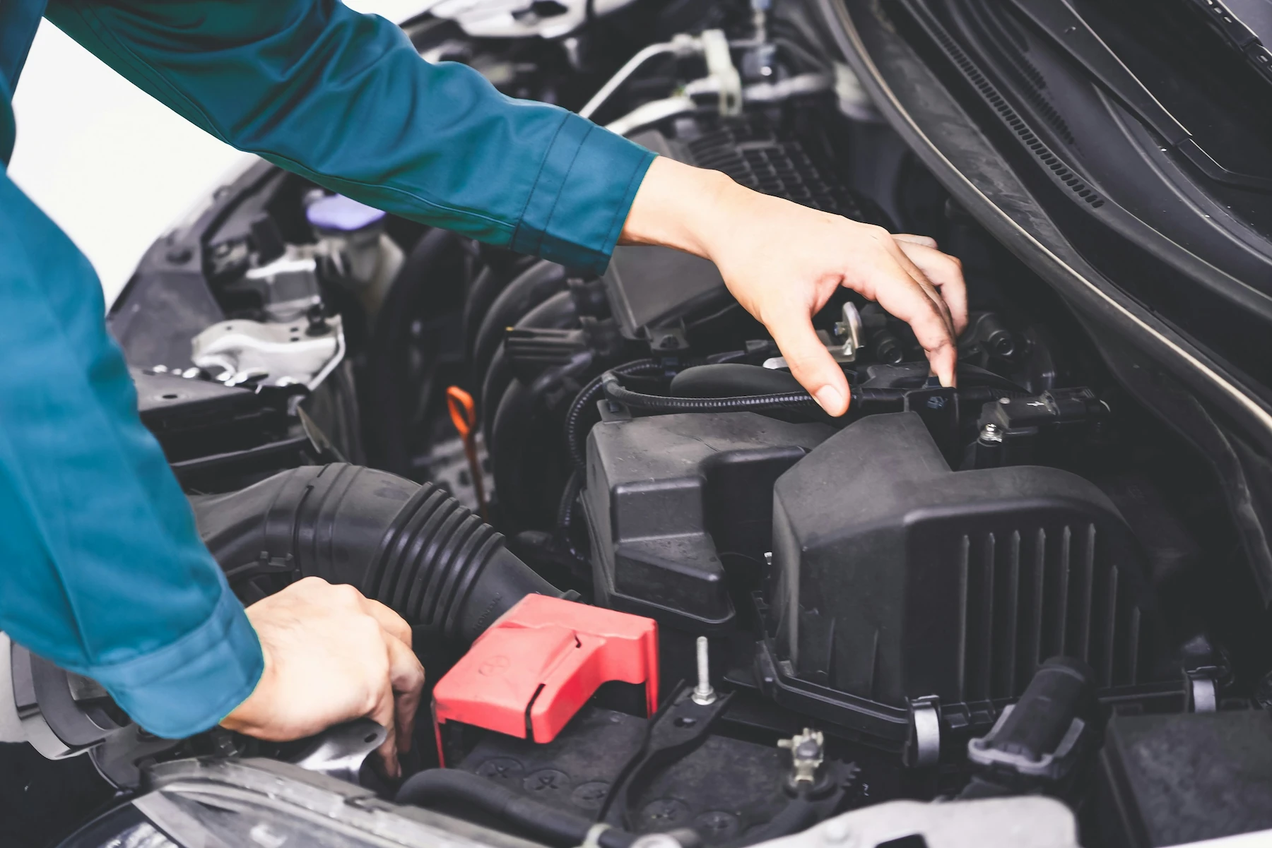 A technician's hands working in a vehicle's engine bay near the air intake housing and battery, with a red terminal cover visible.