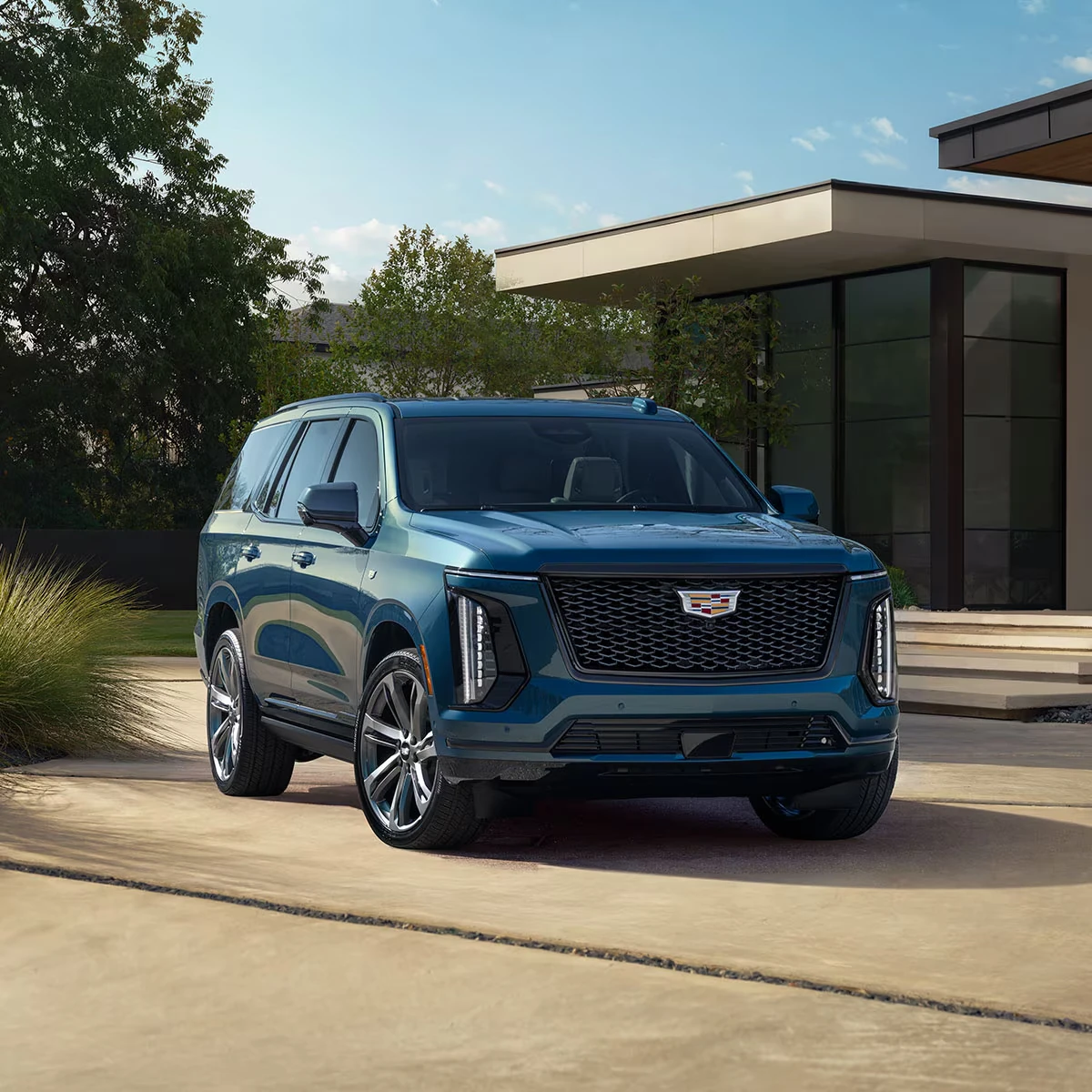 Blue 2027 Cadillac Escalade SUV parked in a driveway in front of a modern house with large windows