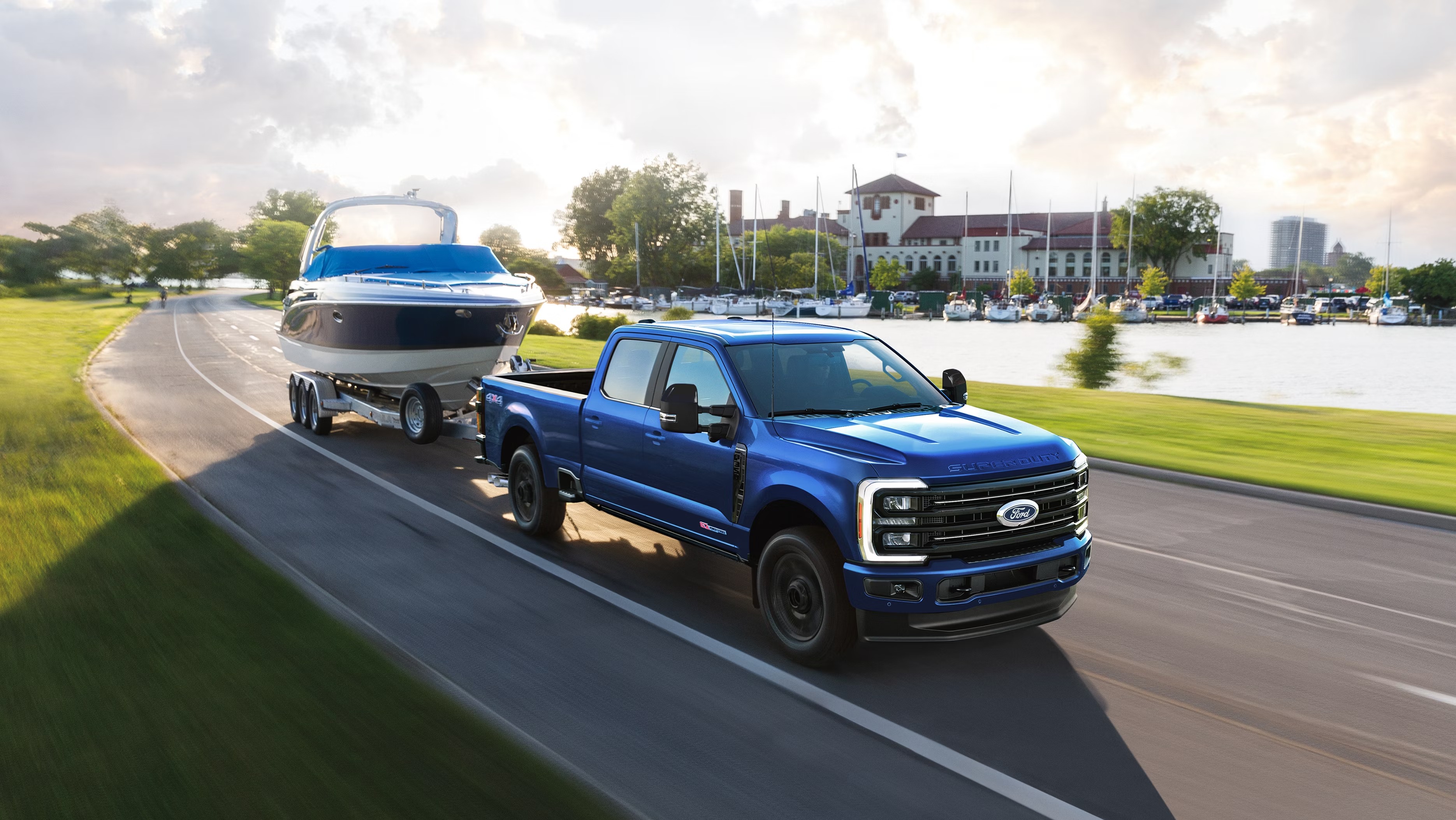 Neptune Blue 2027 Ford F-350 Super Duty Platinum crew cab pickup towing a boat on a trailer along a lakeside road past a marina, shown from a front three-quarter angle.