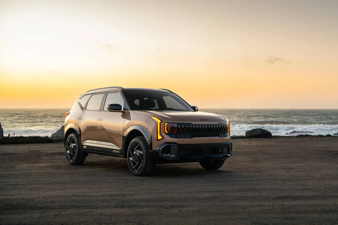 Bronze 2027 Kia Seltos parked on rocks at a coastal overlook, with ocean waves and a sunset sky in the background.