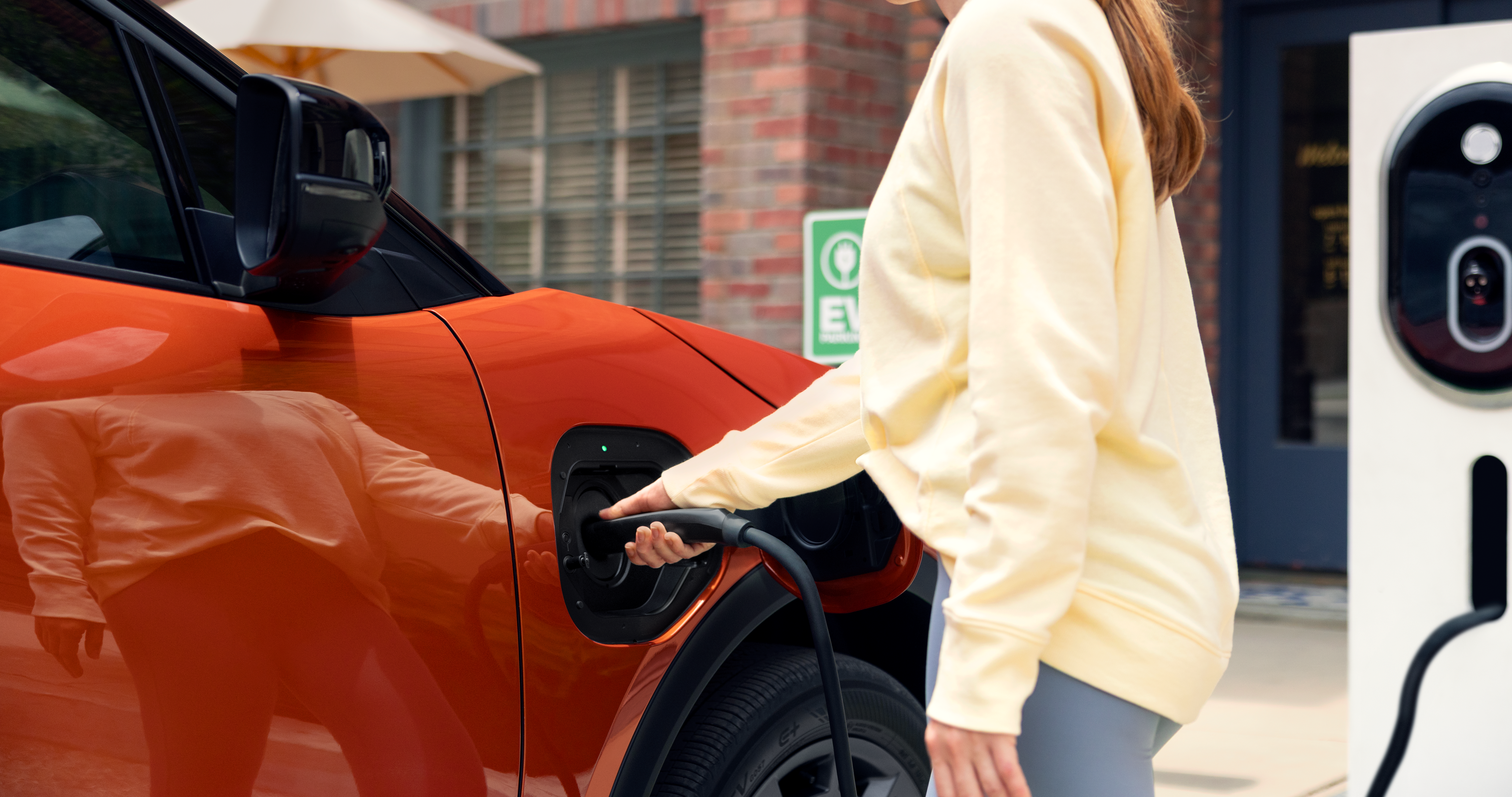 A static close-up image of a person plugging a charging cable into the front driver‑side charging port of an orange Subaru Uncharted. 