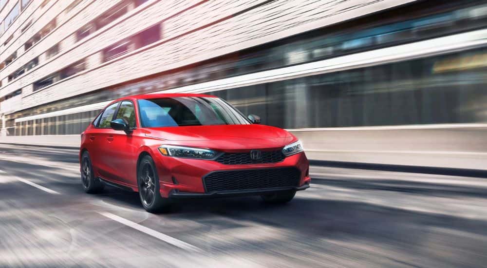 Red 2025 Honda Civic Hybrid Touring driving past blurred buildings.