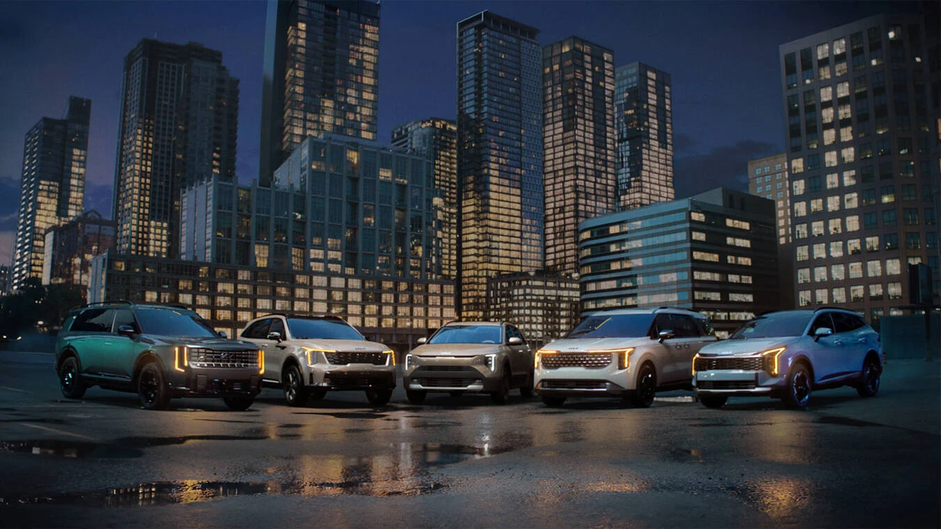 Five Kia SUVs and utility vehicles, including the 2027 Telluride HEV, Sportage HEV, and Carnival HEV, parked together at night in a city lot with amber accent lighting and a lit skyline behind them.