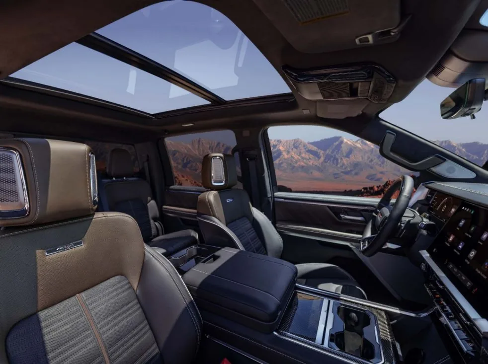 Rear interior view of a GMC pickup truck's two-tone leather captain's chairs and expansive panoramic sunroof, with desert mountains visible through the windows