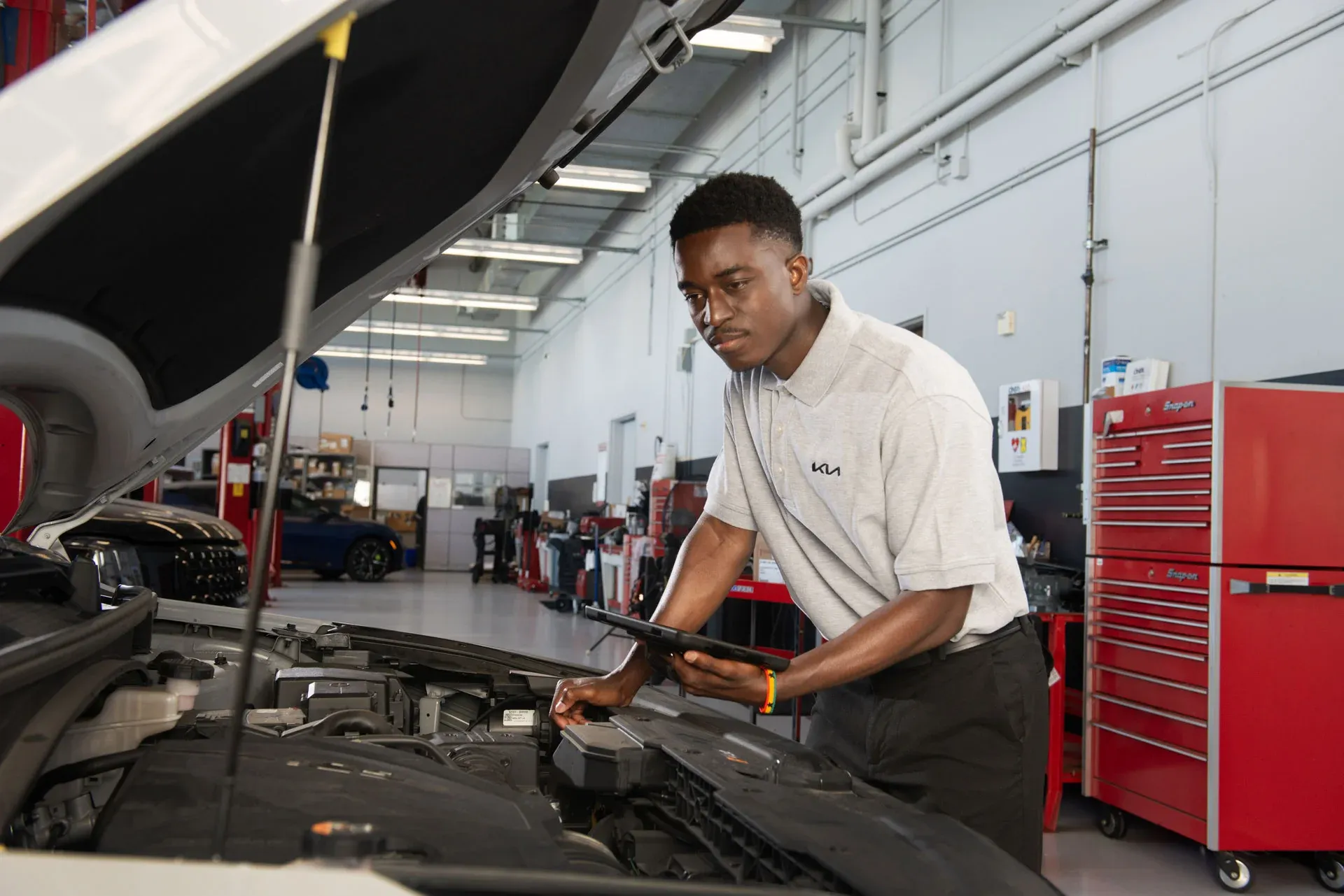 Kia service technician in uniform reviewing a tablet while inspecting a vehicle with the hood open in the service bay