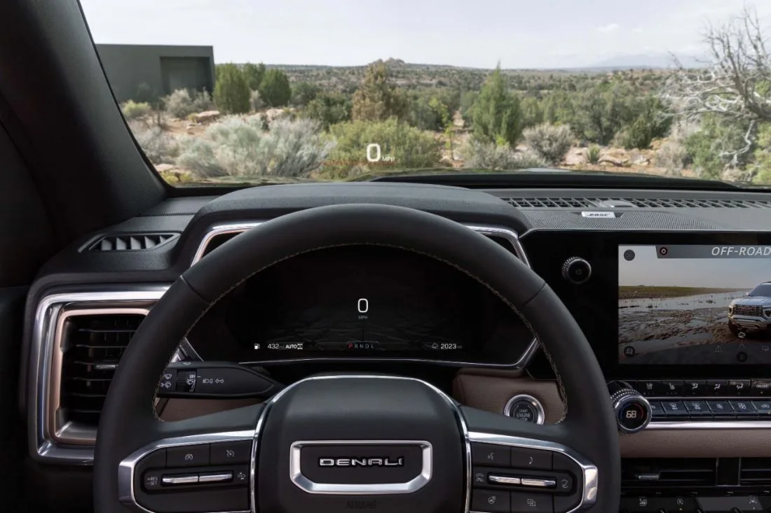 Interior view of a GMC pickup truck with a Denali-badged steering wheel, showing a heads-up display reading 0 mph and "Off-Road" drive mode selected on the center touchscreen, with a desert canyon visible through the windshield