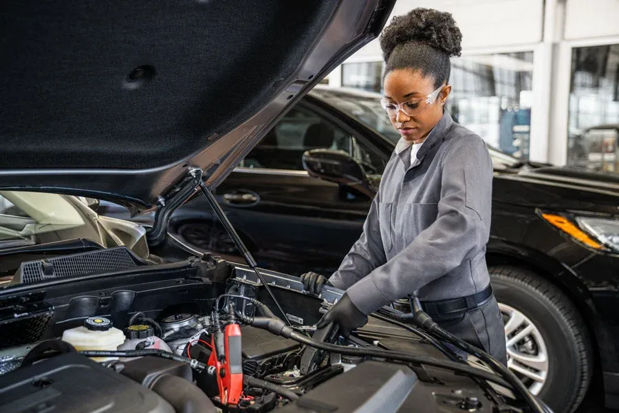 Female technician wearing safety glasses connects jumper cables under an open hood in a dealership service bay, with another vehicle visible in the background