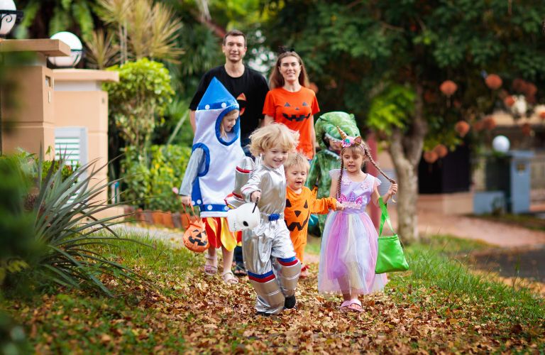 Kids in Costumes Running on a Sidewalk to Trick or Treat with Parents Behind