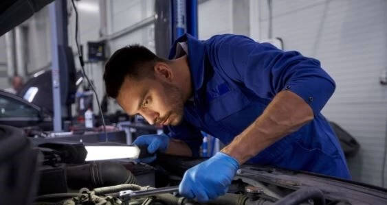 Mechanic in blue overalls and blue latex gloves bent over a car and applying a wrench to a part on the car he is working on