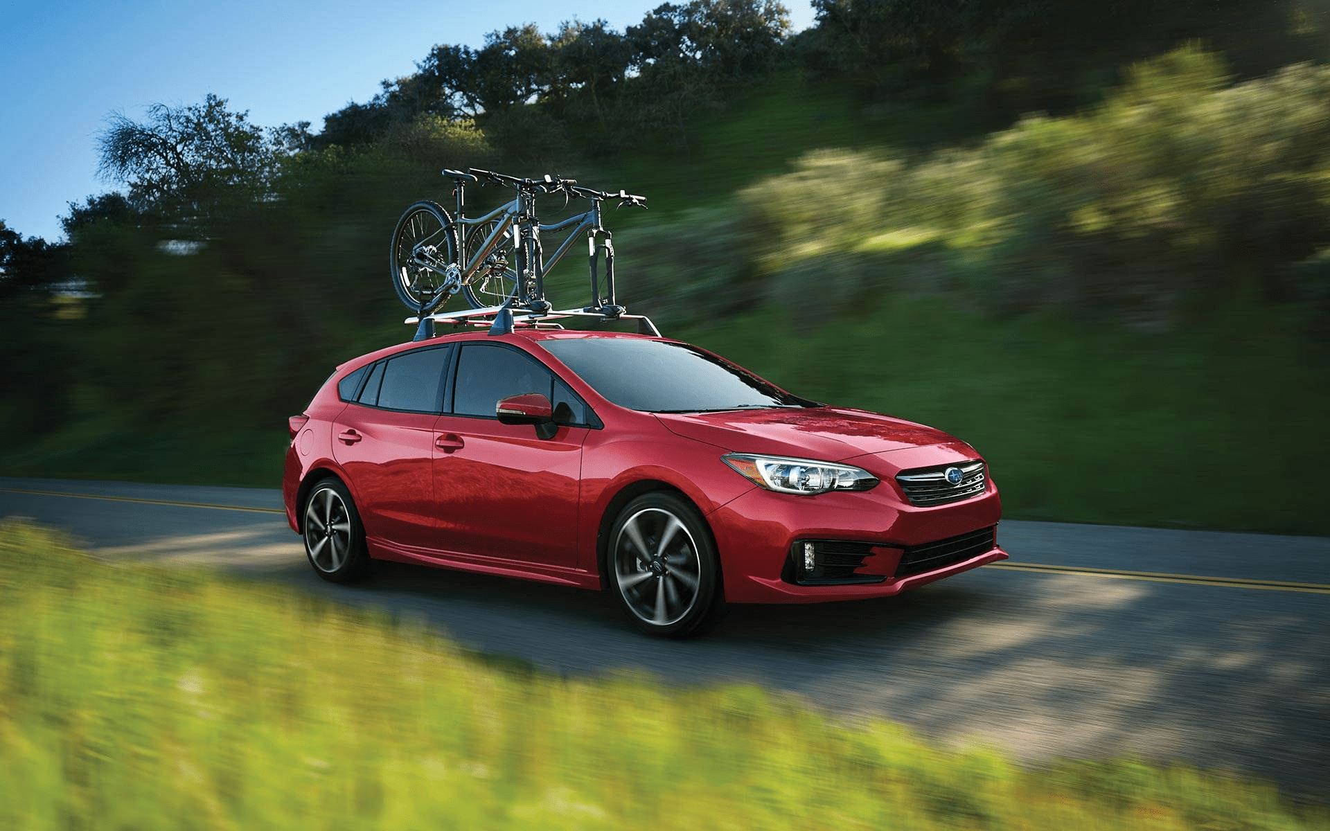 Red Subaru Impreza hatchback with a bike on the roof rack driving along a green hillside road