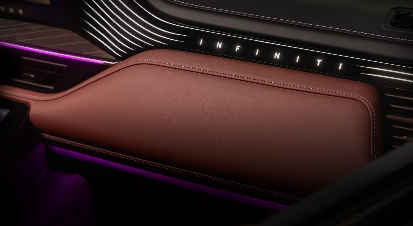 Close-up of the 2027 INFINITI QX80 passenger dashboard with Sepia Brown leather, an illuminated INFINITI logo, and purple ambient light in the footwell.