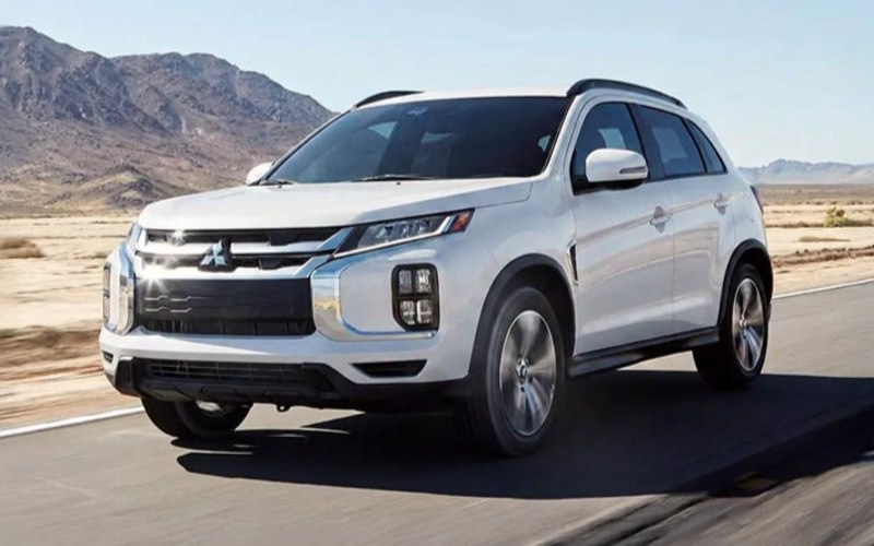 White Mitsubishi Outlander Sport SUV driving along a desert road with rugged mountains in the background, shown from a front three-quarter angle with motion-blurred surroundings.