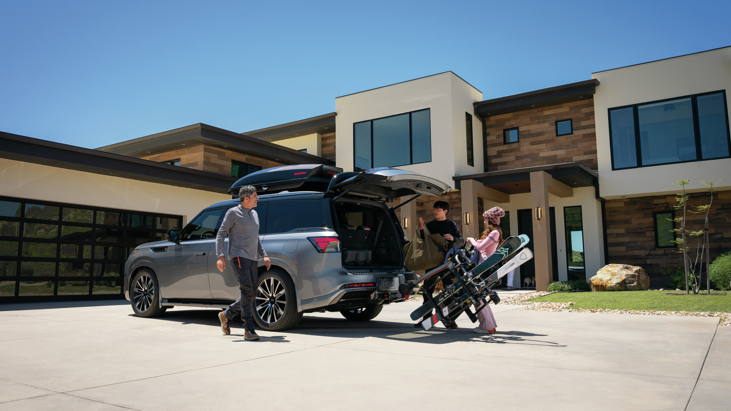 Rear three-quarter view of a gray 2027 INFINITI QX80 with a roof cargo box, hitch-mounted ski rack, and family loading gear into the open cargo area.