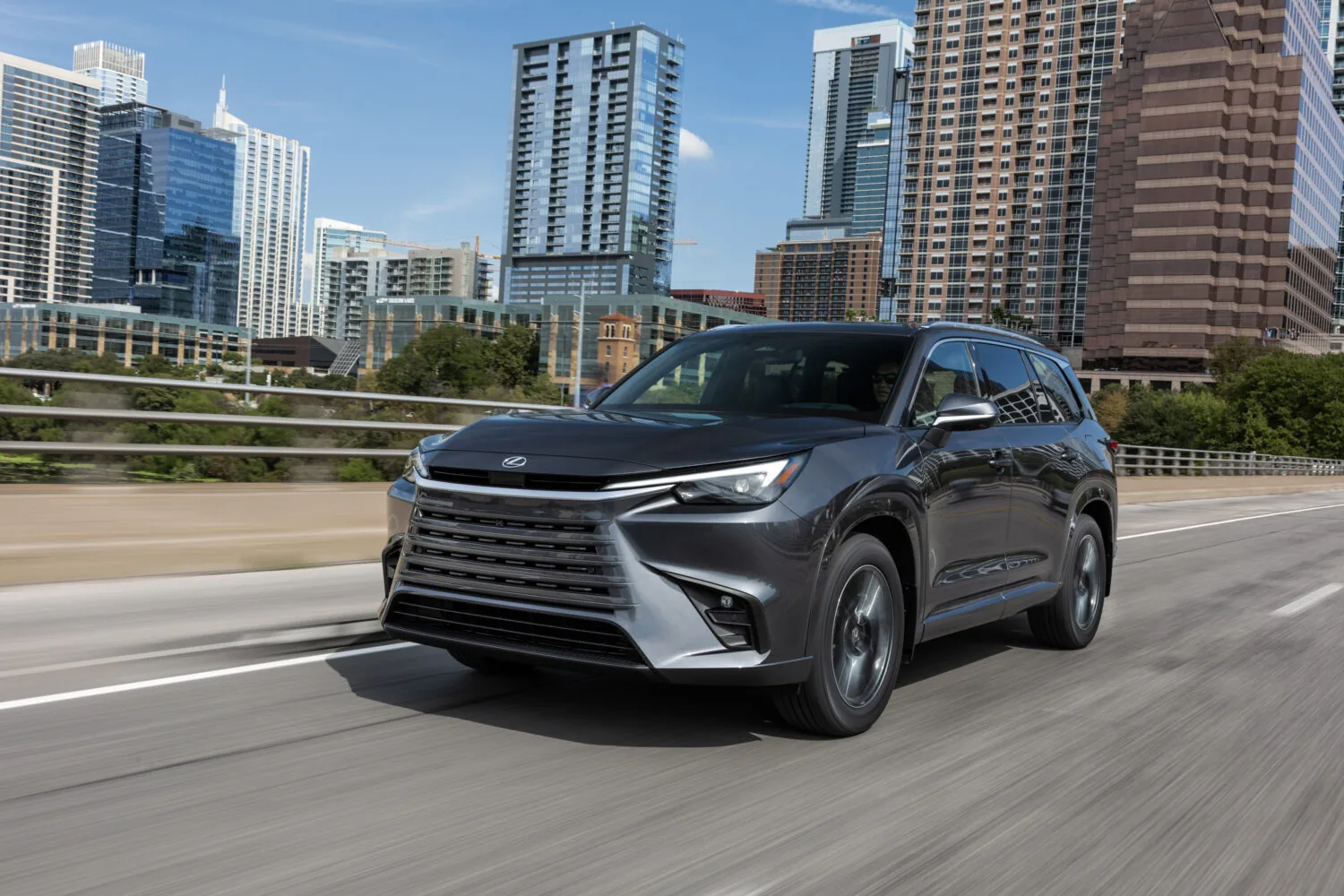 Point-of-view from the driver's seat of a Lexus showing hands on the steering wheel with the Lexus logo, a digital speedometer reading 60 mph, and an infotainment screen displaying a music playlist, with a sunset over a winding road ahead.